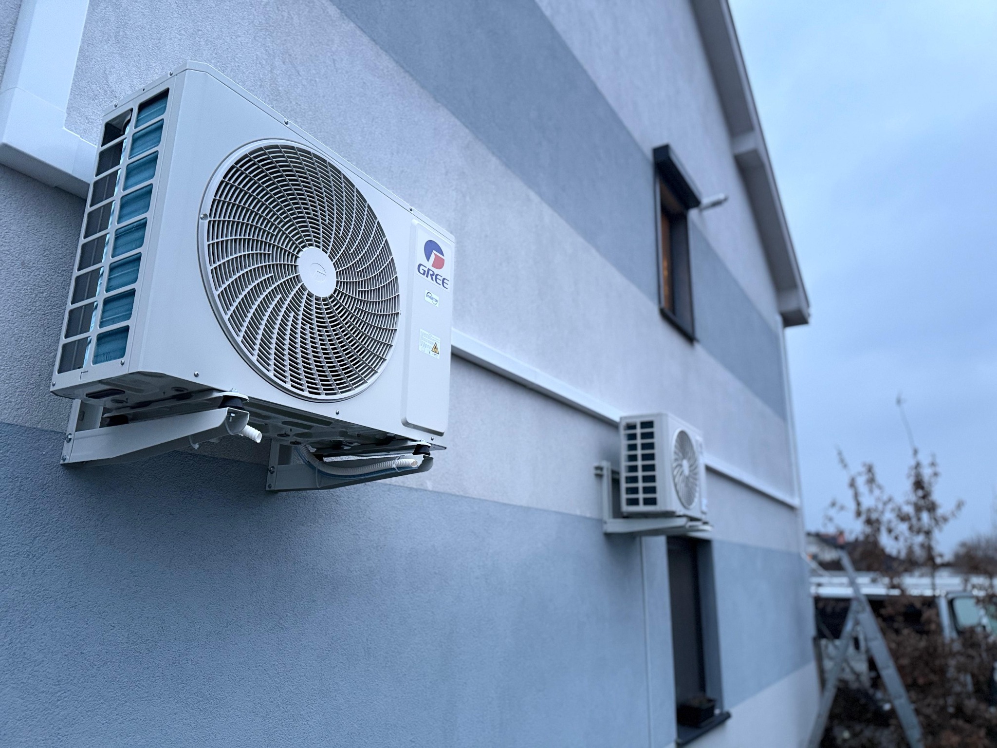 Dwa klimatyzatory marki Gree zamontowane na elewacji budynku z poziomymi pasami w odcieniach szarości. Jeden klimatyzator większy, drugi mniejszy. Na drugim planie drabina i samochód dostawczy.