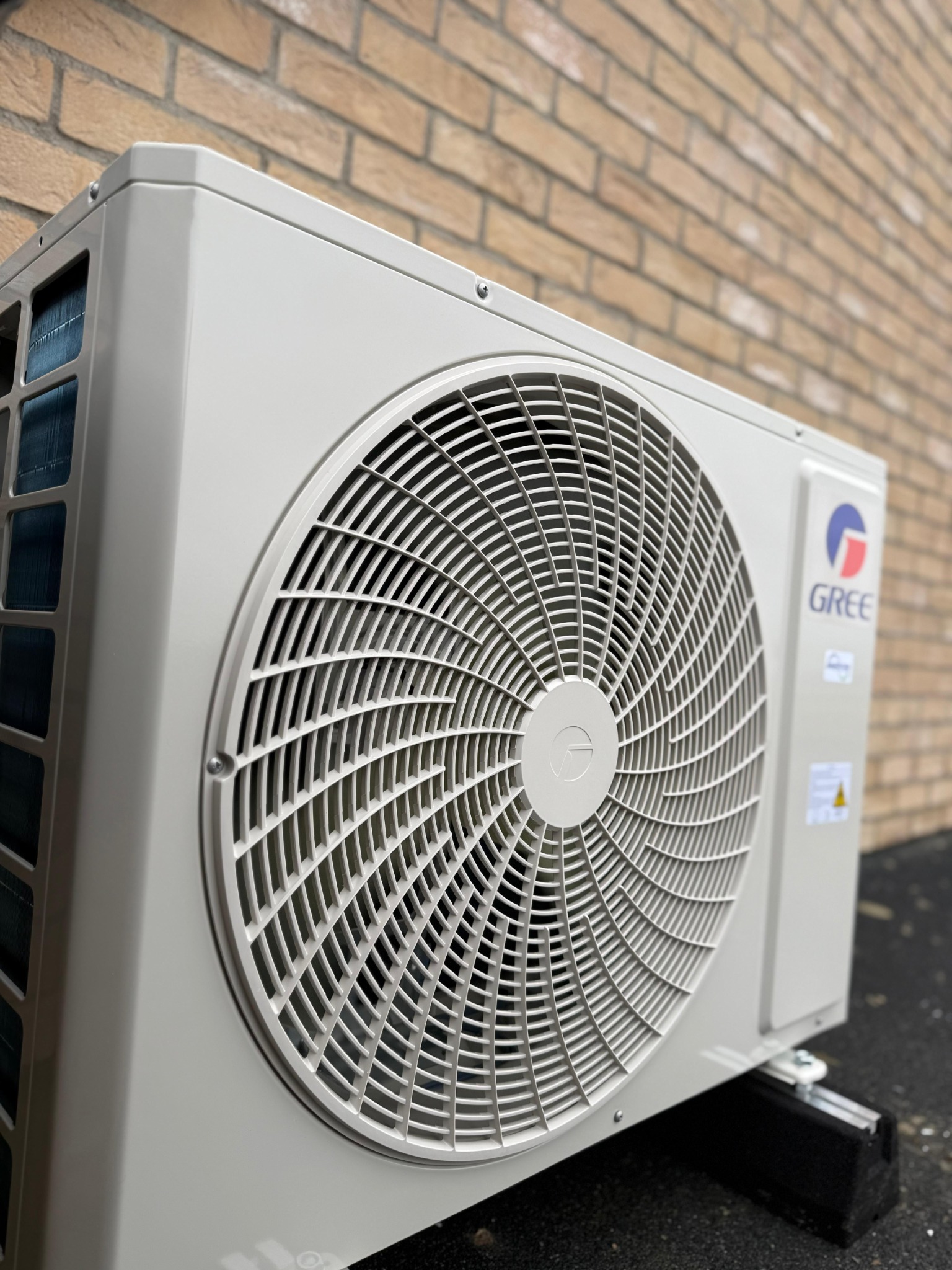 Zewnętrzna jednostka klimatyzacji marki Gree zamontowana na wspornikach, na tle ściany z cegły klinkierowej, widok z dołu.