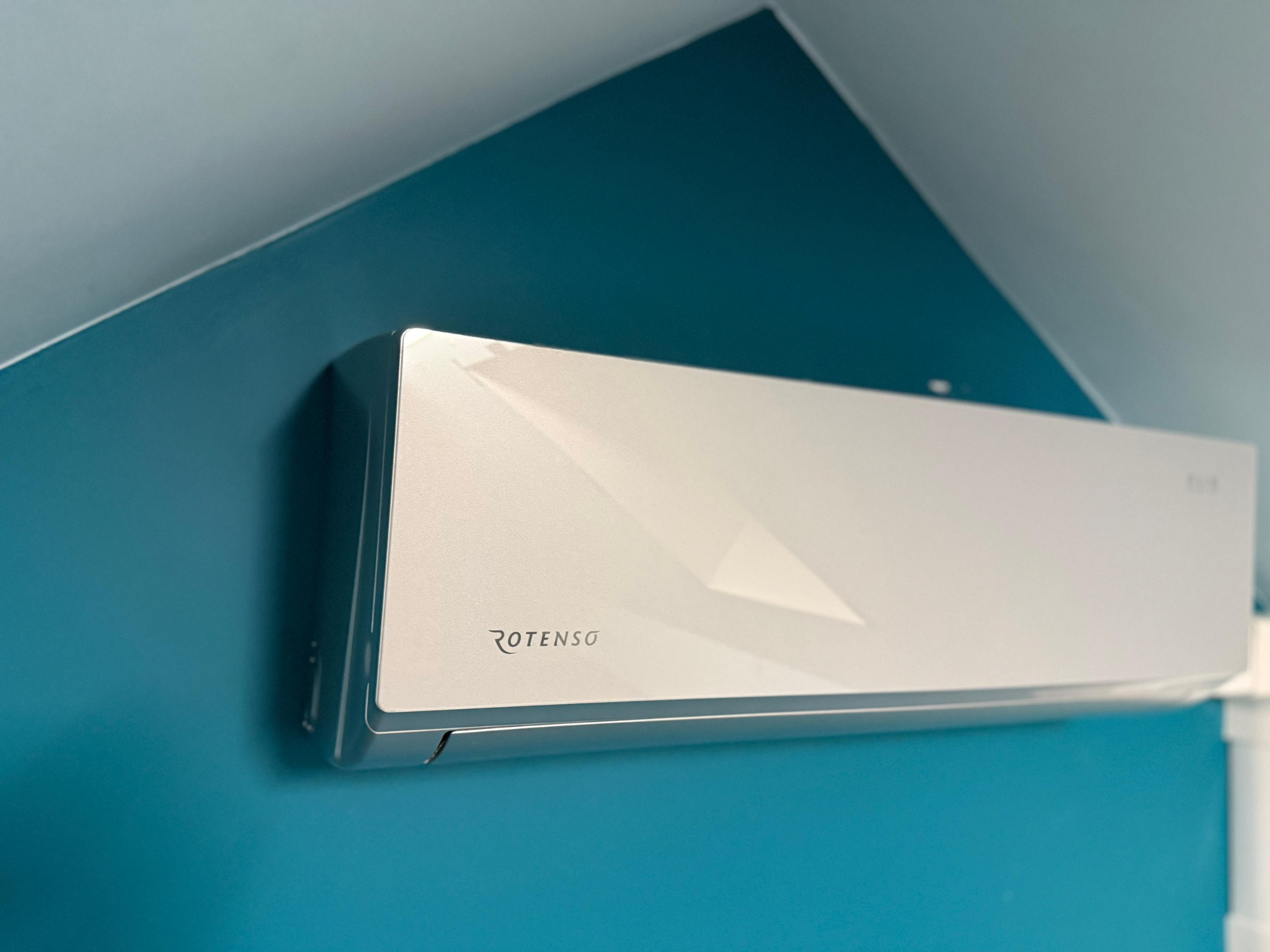 Biała jednostka wewnętrzna klimatyzatora Rotenso zamontowana na turkusowej ścianie w rogu pomieszczenia.