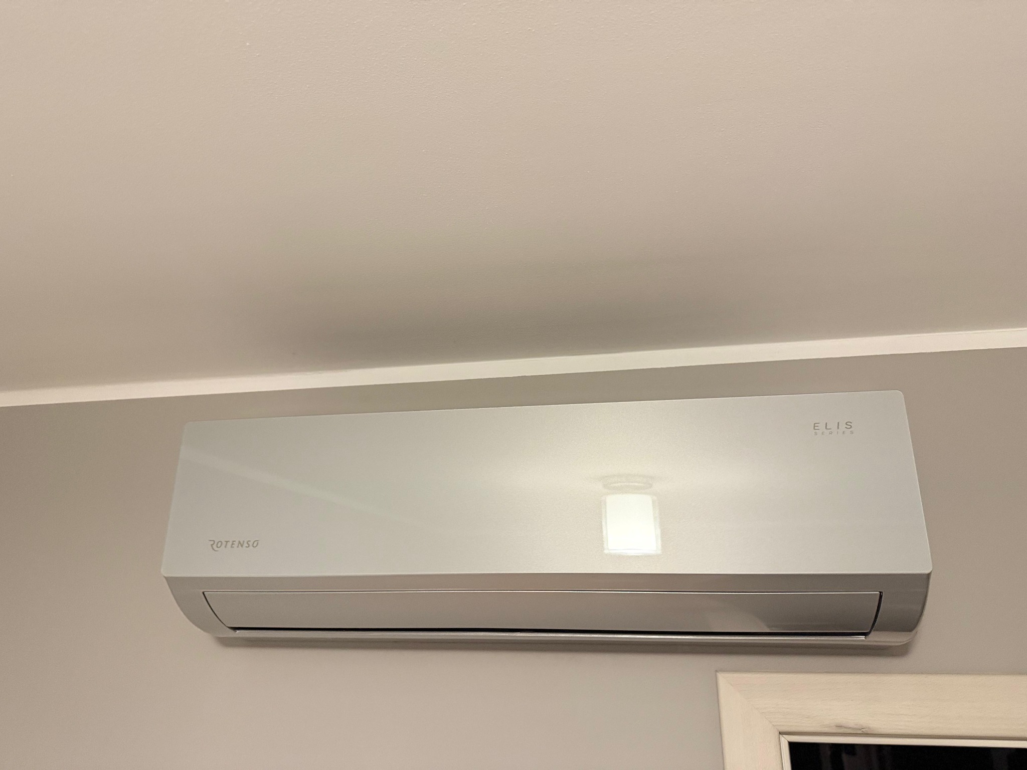 Biała jednostka klimatyzacji Rotenso Elis Series zamontowana na ścianie w jasnym pomieszczeniu, widoczny fragment ramy okiennej w prawym dolnym rogu.