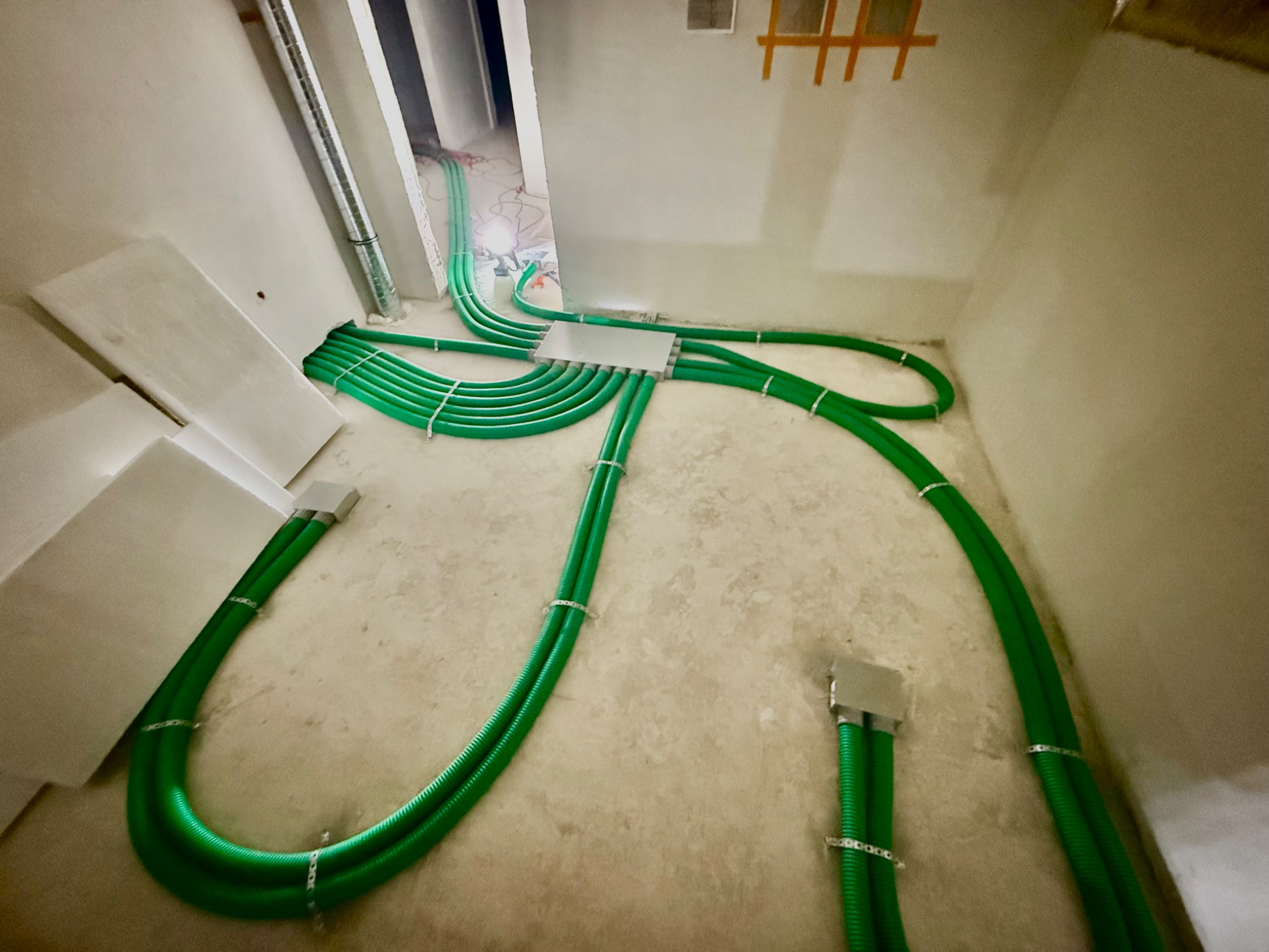 Zielone rury wentylacyjne ułożone na podłodze w surowym wnętrzu, połączone w centralnym punkcie. Instalacja przed zakryciem, widoczne uchwyty mocujące.