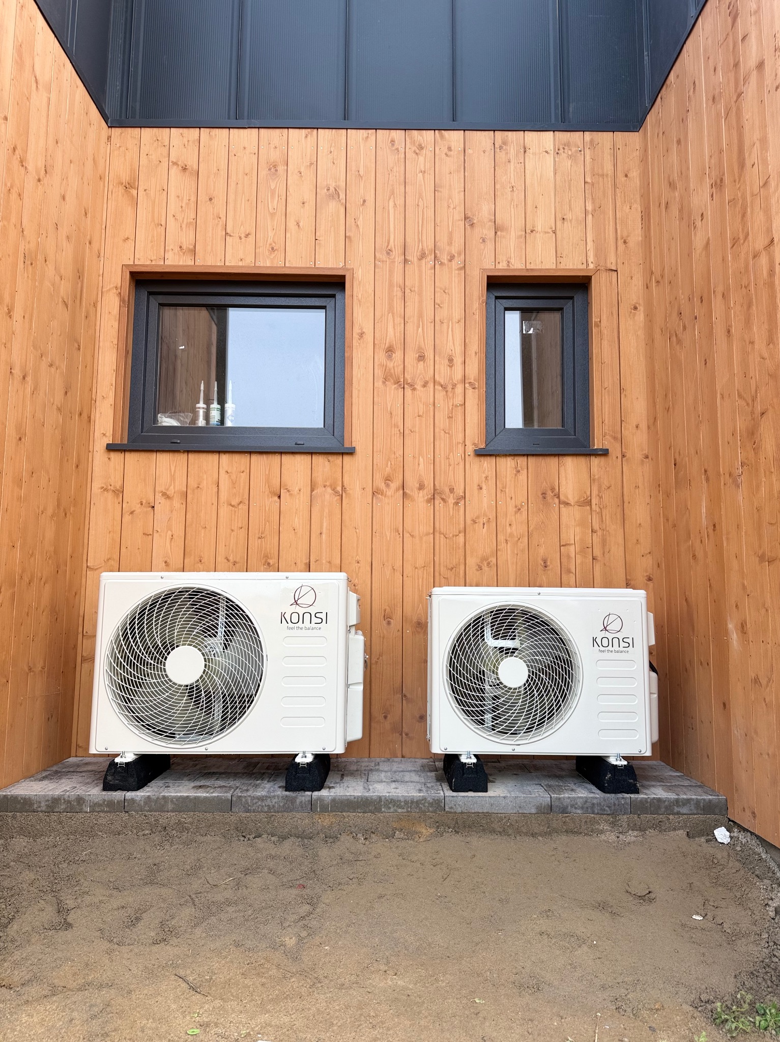 Dwa białe klimatyzatory marki Konsi zamontowane na zewnątrz drewnianego budynku z dwoma oknami o różnych rozmiarach, na kostce brukowej.