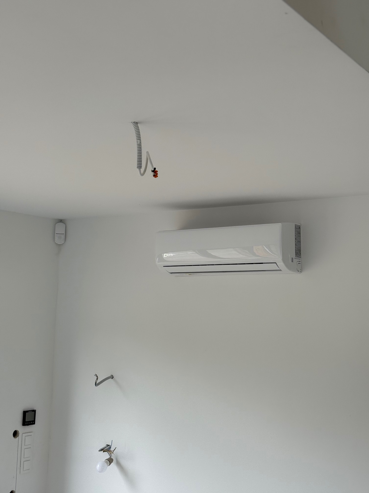 Nowoczesny, biały klimatyzator KONSi zamontowany na białej ścianie w minimalistycznym wnętrzu. Widoczne elementy instalacji elektrycznej.