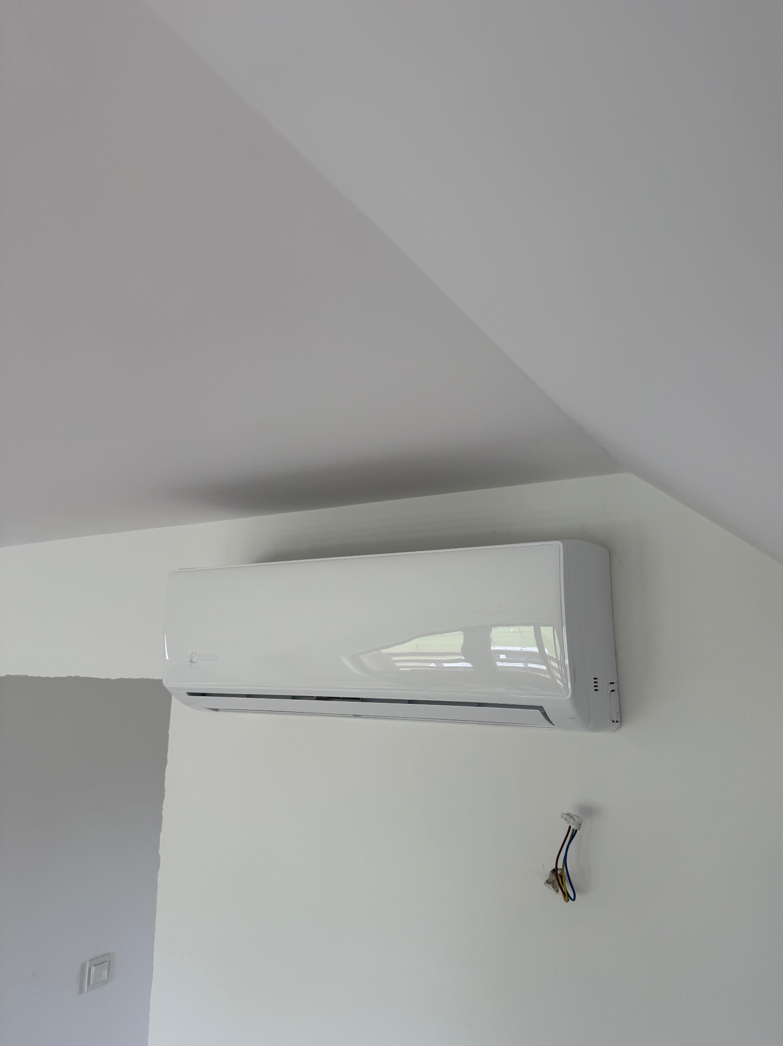 Biały klimatyzator marki Konsi zamontowany na białej ścianie pod skosem dachu, z widocznymi przewodami instalacyjnymi, sugerujący montaż w trakcie wykończenia wnętrza.