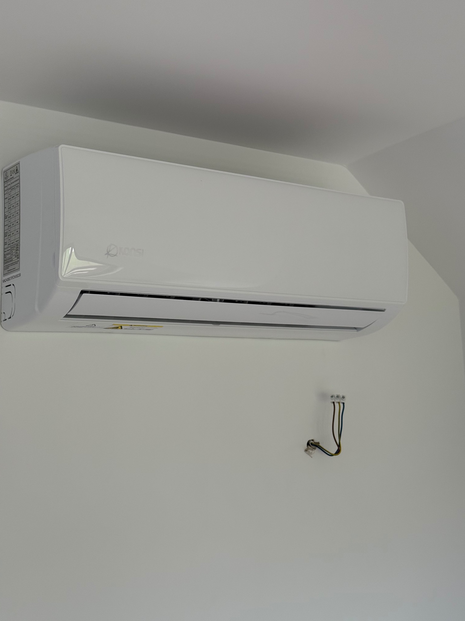 Biała klimatyzacja marki Konsi zamontowana na ścianie, widoczne wystające przewody elektryczne, minimalistyczny design, wnętrze w jasnych barwach.