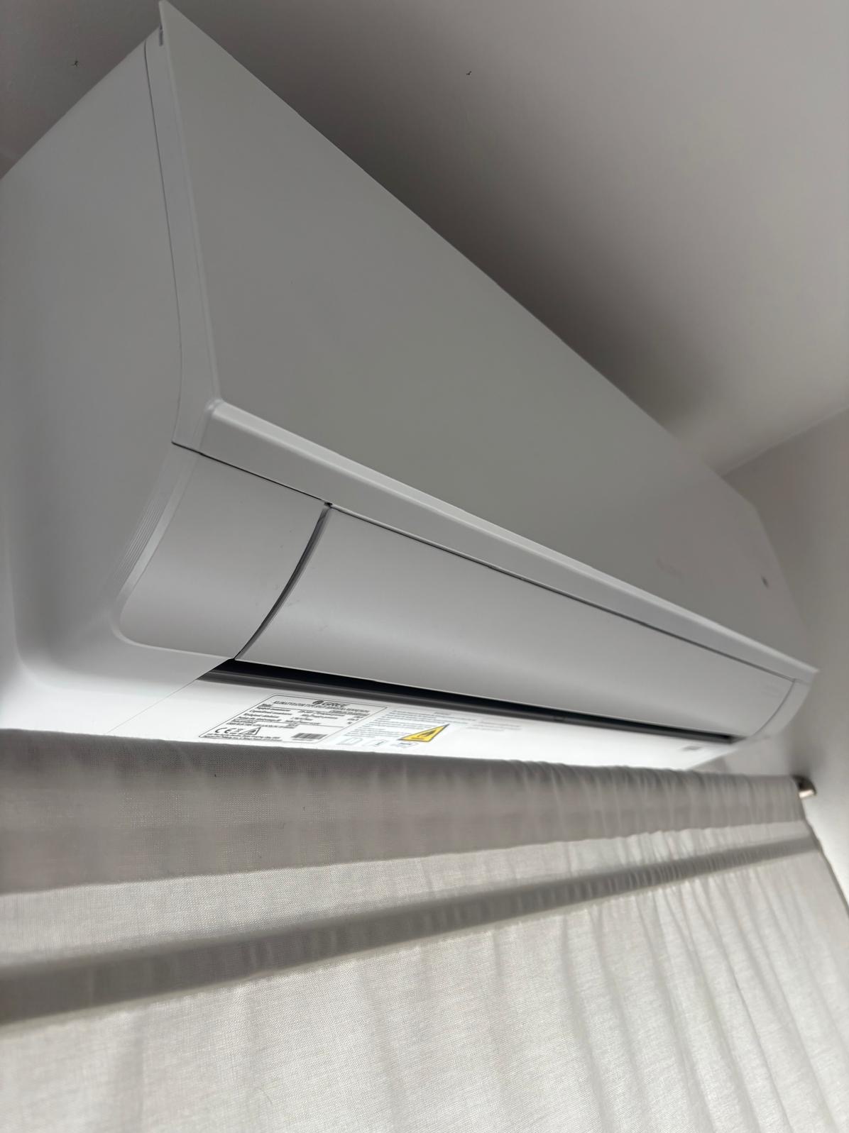 Biały klimatyzator ścienny, widoczny od dołu, zamontowany w pomieszczeniu z zasłonami. Widoczne naklejki informacyjne na urządzeniu.