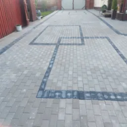 Jasnoszara kostka brukowa z ciemnymi obramowaniami tworzącymi kwadratowe wzory na podjeździe. Widoczny fragment ogrodzenia i brama w tle.