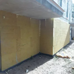 Ocieplenie zewnętrzne budynku z widoczną wełną mineralną, zabezpieczoną folią fundamentową przy gruncie, z oknem zabezpieczonym folią ochronną.