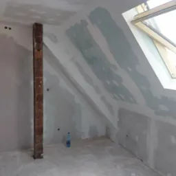 Poddasze w trakcie prac wykończeniowych: ściany i skosy obłożone płytami kartonowo-gipsowymi, częściowo zaszpachlowane, widoczny drewniany słup, okno dachowe i butelka wody na podłodze.