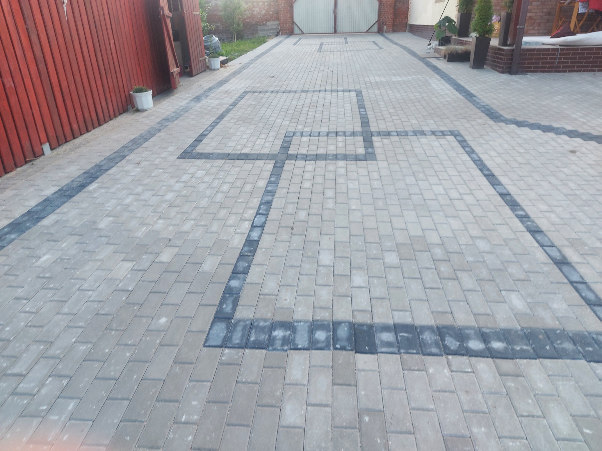 Jasnoszara kostka brukowa z ciemnymi obramowaniami tworzącymi kwadratowe wzory na podjeździe. Widoczny fragment ogrodzenia i brama w tle.
