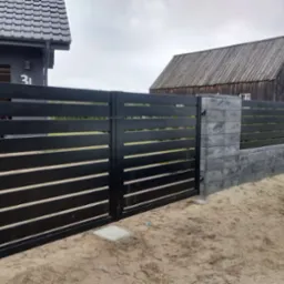 Nowoczesna czarna brama wjazdowa z poziomymi panelami i fragment ogrodzenia panelowego z szarej kostki betonowej na tle budynków.