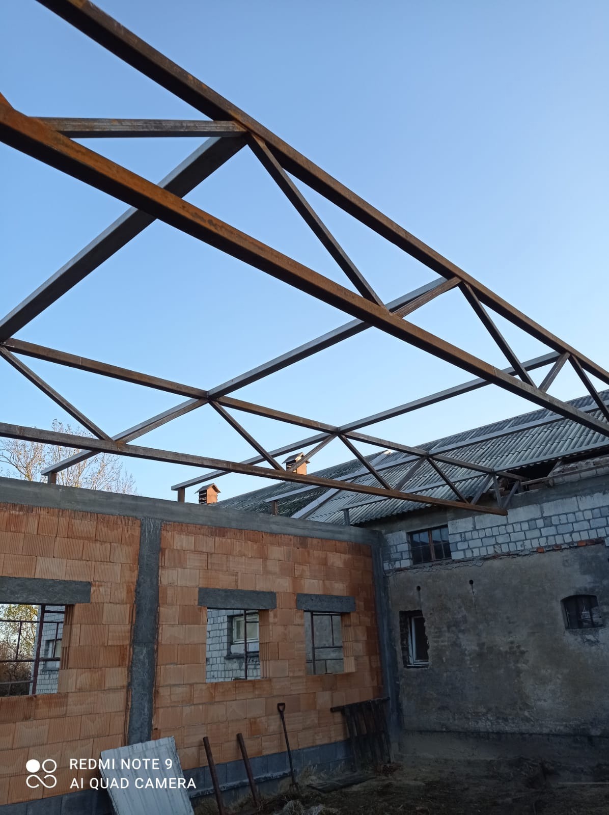 Stalowa konstrukcja dachu w trakcie montażu, widoczne mury z cegły kratówki i fragment starego budynku w tle, błękitne niebo.