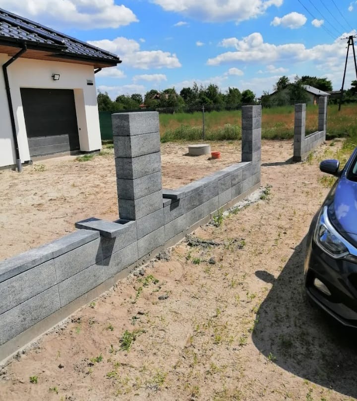 Wznoszenie niskiego, szarego murku z betonowych bloczków na posesji, widoczne słupki i fragment zaparkowanego samochodu.