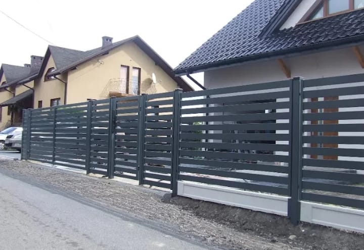 Szare ogrodzenie panelowe z poziomych listew, betonowa podmurówka, widok z chodnika na tle domów.