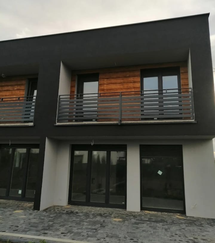 Nowoczesny budynek dwukondygnacyjny z ciemnoszarą elewacją, drewnianymi panelami, szarymi balkonami z poziomymi barierkami i przeszklonymi drzwiami na parterze, wyłożony kostką brukową.