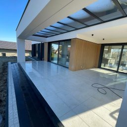 Frączek Firma Budowlana - Nowoczesny taras z jasnymi płytkami i czarną kostką obok, zadaszony szklaną konstrukcją, w tle dom z dużymi oknami przesuwnymi. Słoneczny dzień.