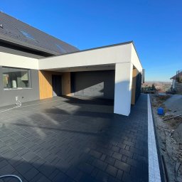 Frączek Firma Budowlana - Nowoczesny podjazd z ciemnej kostki brukowej przy domu z garażem, wykończony białą obwódką. Błękitne niebo w tle.