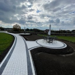 Frączek Firma Budowlana - Ścieżka z kostki brukowej w ogrodzie z okrągłym patio i meblami ogrodowymi pod parasolem. Widok na pochmurne niebo i drzewa w tle.