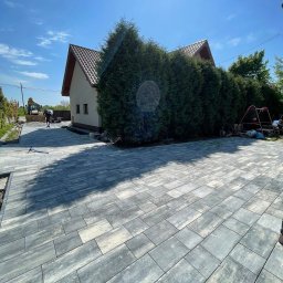 Frączek Firma Budowlana - Ułożona, szara kostka brukowa na podjeździe przed domem z zielonymi tujami. W tle widoczny sprzęt budowlany i pracownik. Słoneczny dzień.