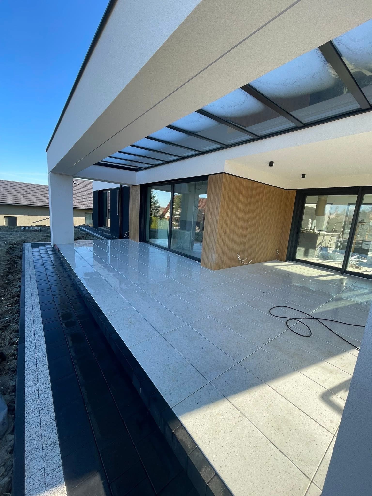 Nowoczesny taras z jasnymi płytkami i czarną kostką obok, zadaszony szklaną konstrukcją, w tle dom z dużymi oknami przesuwnymi. Słoneczny dzień.