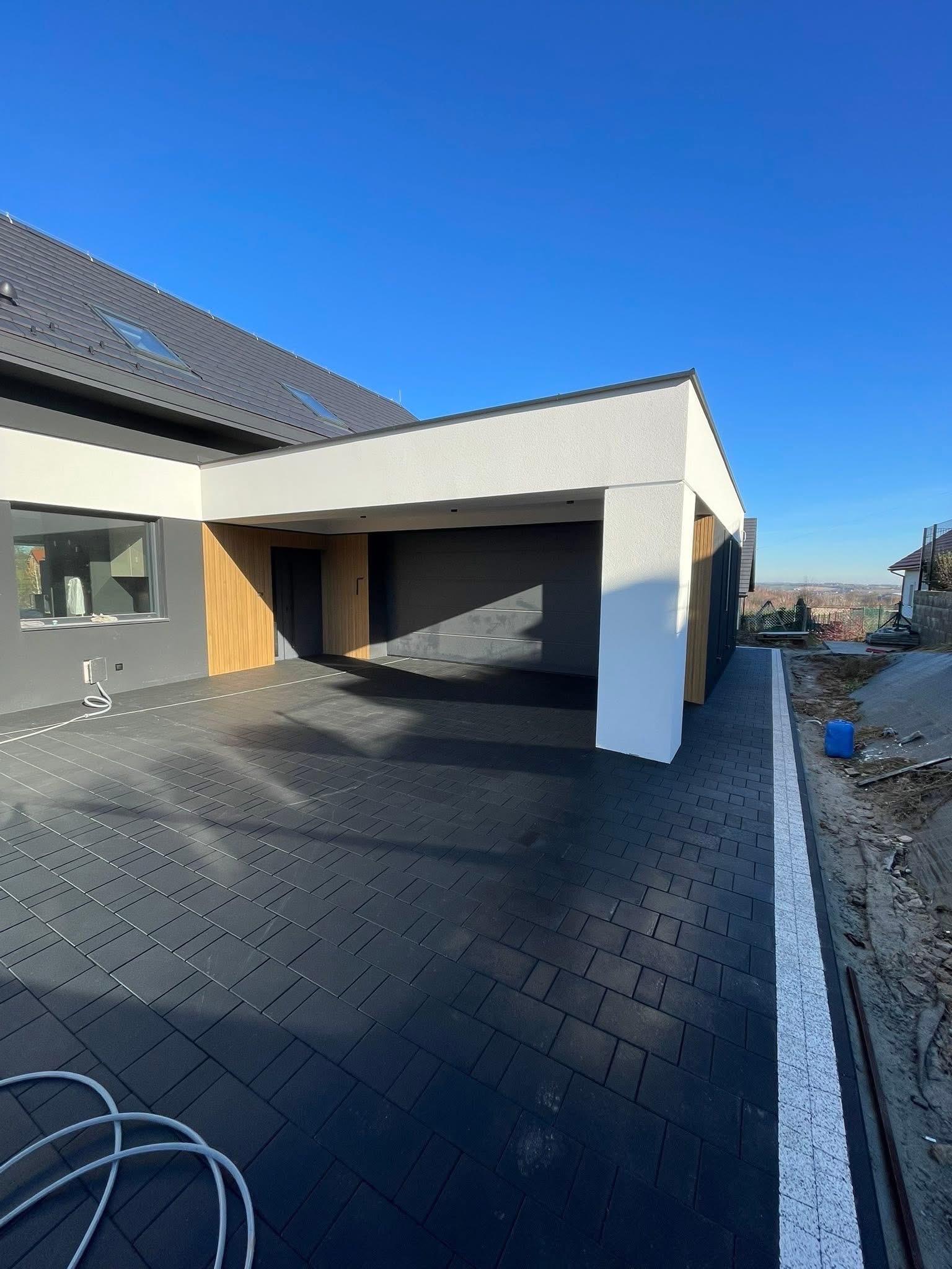 Nowoczesny podjazd z ciemnej kostki brukowej przy domu z garażem, wykończony białą obwódką. Błękitne niebo w tle.