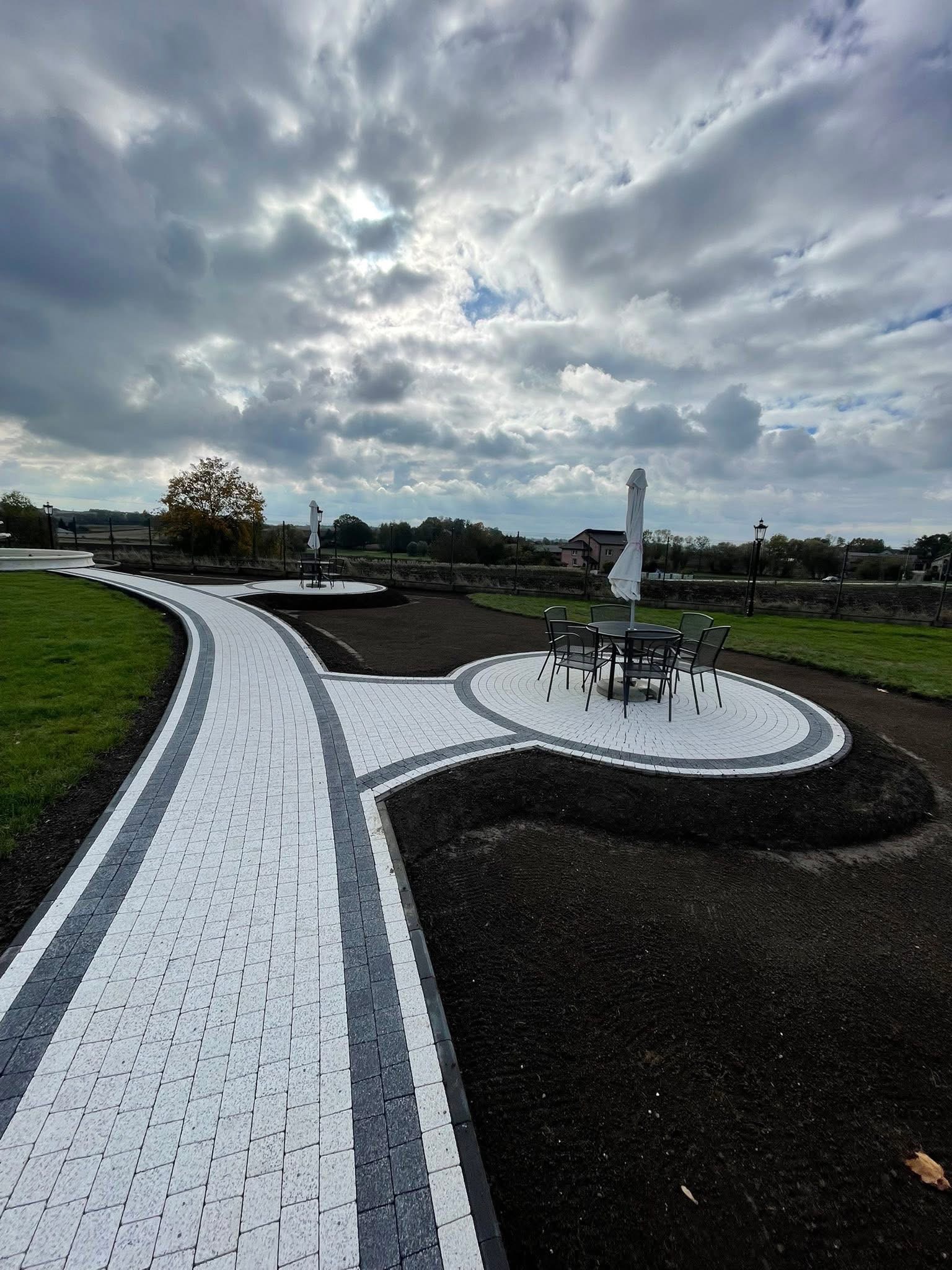 Ścieżka z kostki brukowej w ogrodzie z okrągłym patio i meblami ogrodowymi pod parasolem. Widok na pochmurne niebo i drzewa w tle.