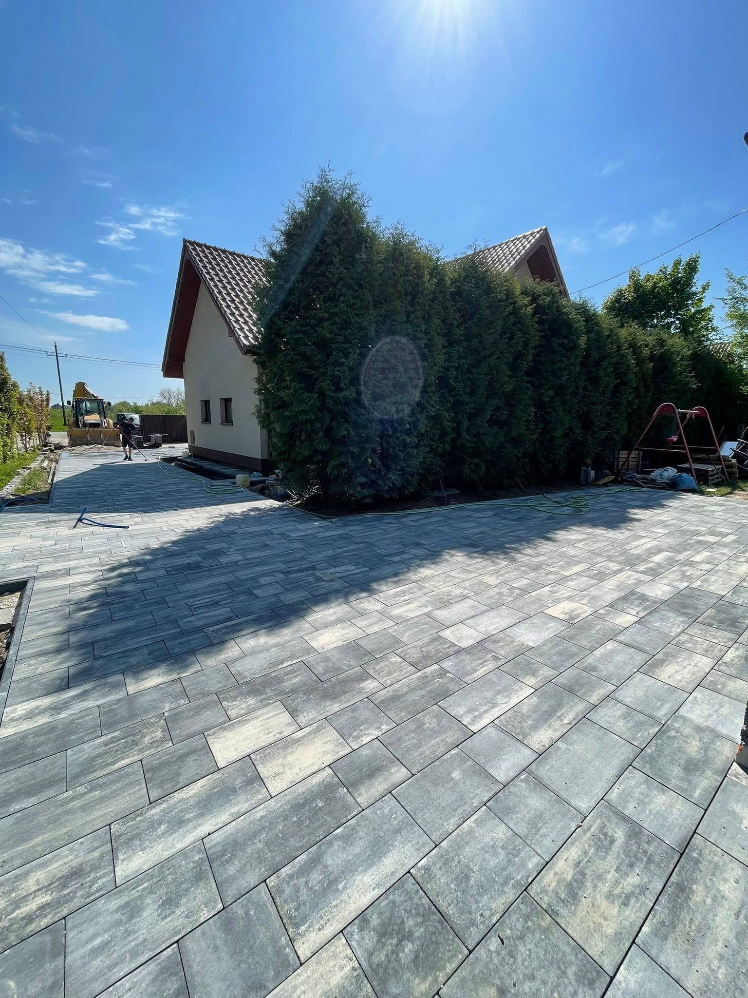 Ułożona, szara kostka brukowa na podjeździe przed domem z zielonymi tujami. W tle widoczny sprzęt budowlany i pracownik. Słoneczny dzień.