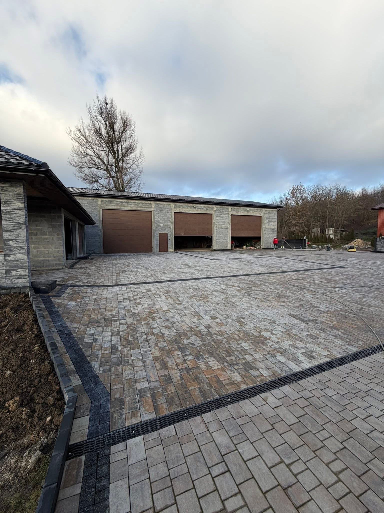 Ukończona nawierzchnia z kostki brukowej w odcieniach brązu i szarości, z czarnym obramowaniem i odpływem liniowym. W tle budynek z bramami garażowymi.