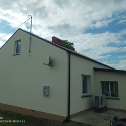 ABC Docieplenia - Jednorodzinny dom po renowacji elewacji w jasnym odcieniu beżu, z ciemnobrązowym cokołem i czarnymi elementami wykończeniowymi dachu oraz rynnami, antena satelitarna na ścianie, w tle błękitne...