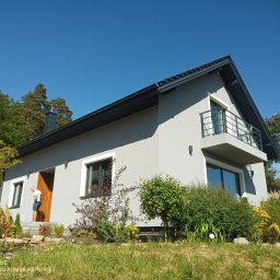 ABC Docieplenia - Nowoczesny dom jednorodzinny z szarą elewacją, balkonem z metalową balustradą i drewnianymi drzwiami wejściowymi, otoczony zielenią i drzewami w słoneczny dzień.