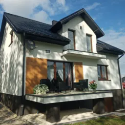 Nowoczesny, dwukondygnacyjny dom z białymi ścianami, ciemnym dachem i drewnianymi akcentami, z tarasem wyposażonym w czarne meble ogrodowe i kwiaty, widok z boku na tle błękitnego nieba z chmurami.