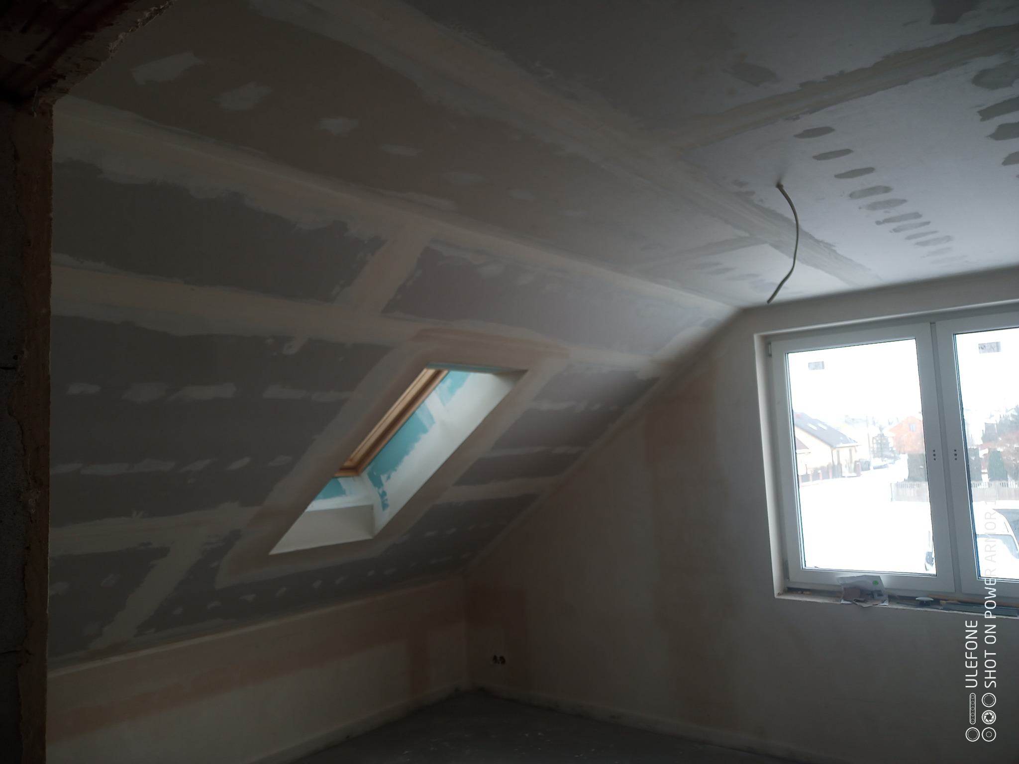 Poddasze z oknem dachowym w trakcie wykańczania płytami gipsowo-kartonowymi, widoczne szpachlowane łączenia płyt, wystający kabel elektryczny z sufitu, widok na zimowy krajobraz za oknem.