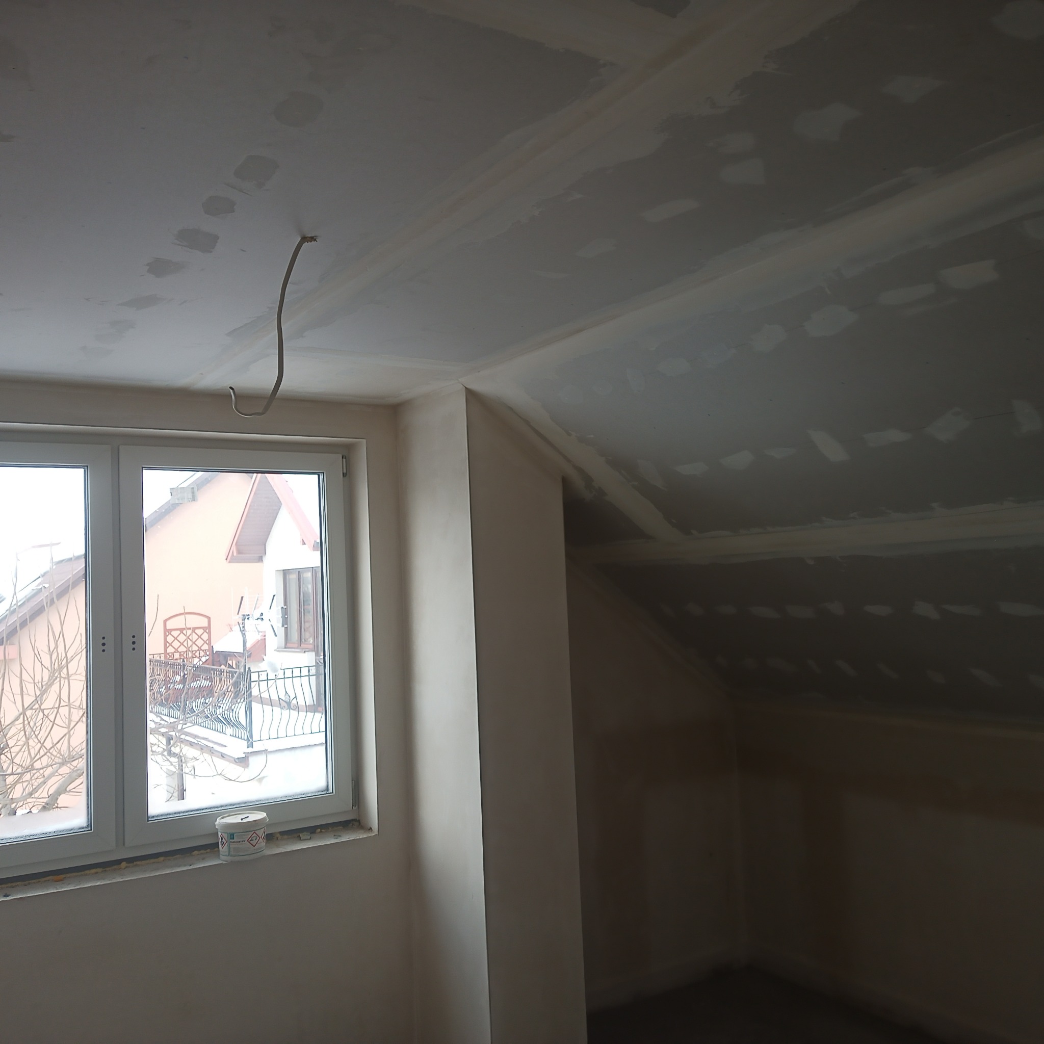Poddasze w trakcie wykańczania płytami kartonowo-gipsowymi, widoczne łączenia szpachlowane, wystający kabel elektryczny i okno z widokiem na zimowy krajobraz.
