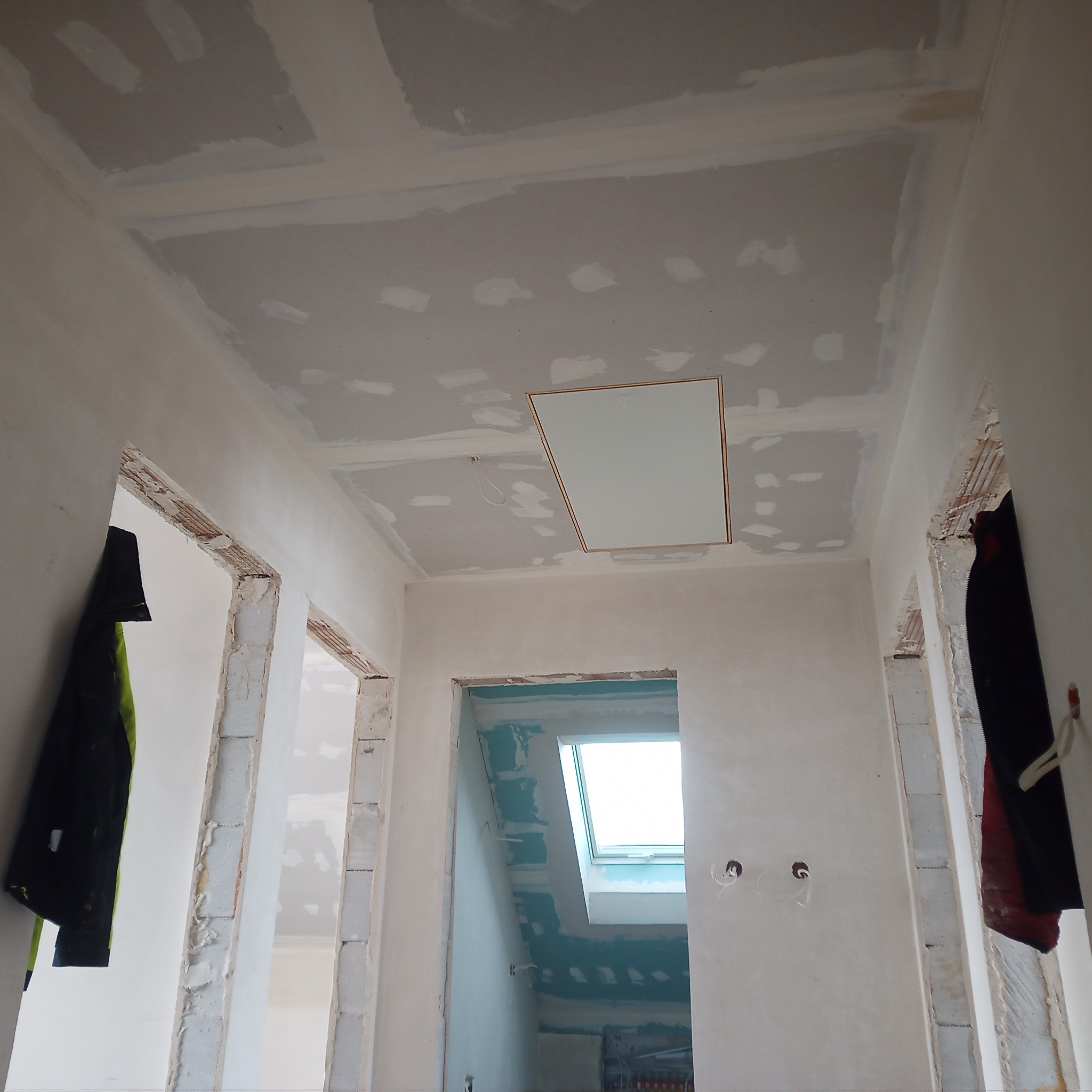 Wnętrze w trakcie remontu: sufit z płyt kartonowo-gipsowych z otworem rewizyjnym, ściany przygotowane do malowania, widoczne fragmenty konstrukcji ścian działowych i okno dachowe.