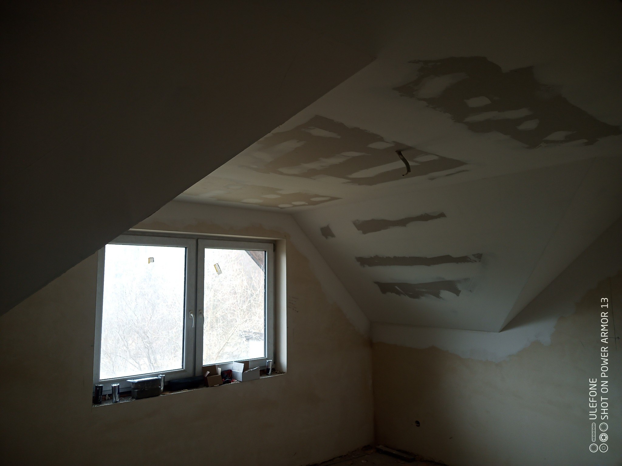 Poddasze w trakcie remontu: okno z widokiem na drzewa, ściany z widocznymi śladami szpachlowania i gładzi, sufit z fragmentami wypełnionymi gipsem.