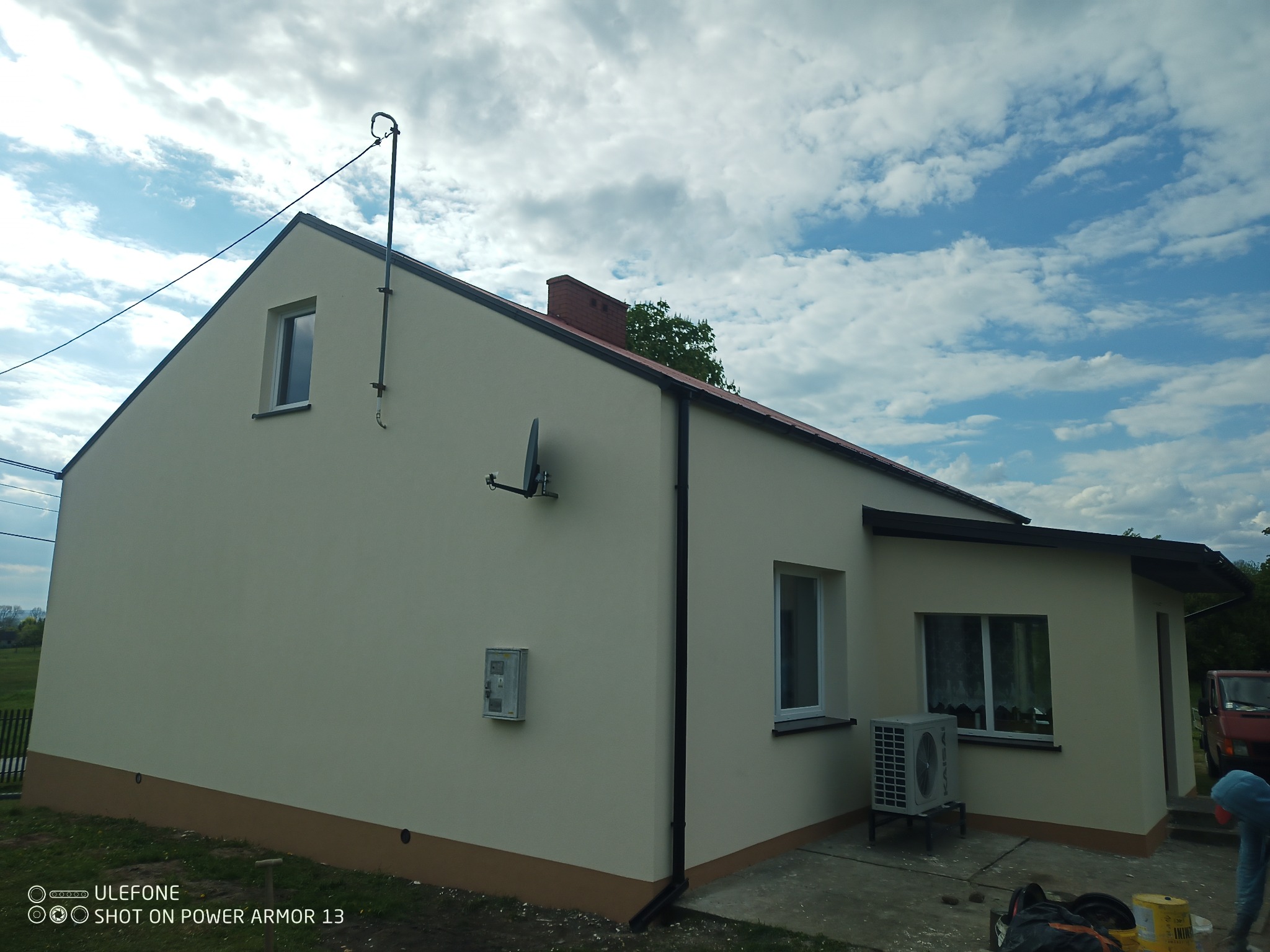 Jednorodzinny dom po renowacji elewacji w jasnym odcieniu beżu, z ciemnobrązowym cokołem i czarnymi elementami wykończeniowymi dachu oraz rynnami, antena satelitarna na ścianie, w tle błękitne...