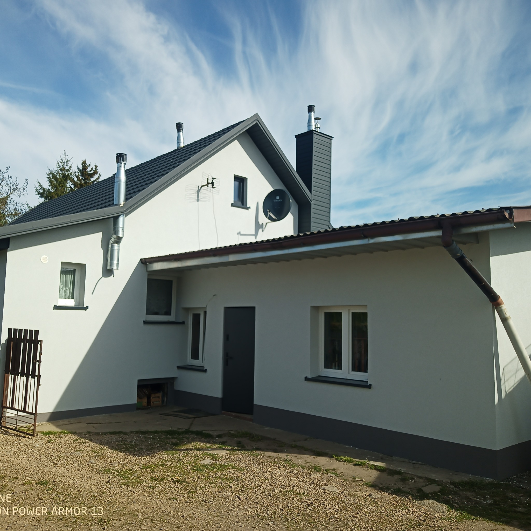 Boczny widok świeżo otynkowanego i pomalowanego na biało domu jednorodzinnego z szarym dachem, brązowymi rynnami i dwoma kominami na tle błękitnego nieba z pierzastymi chmurami.