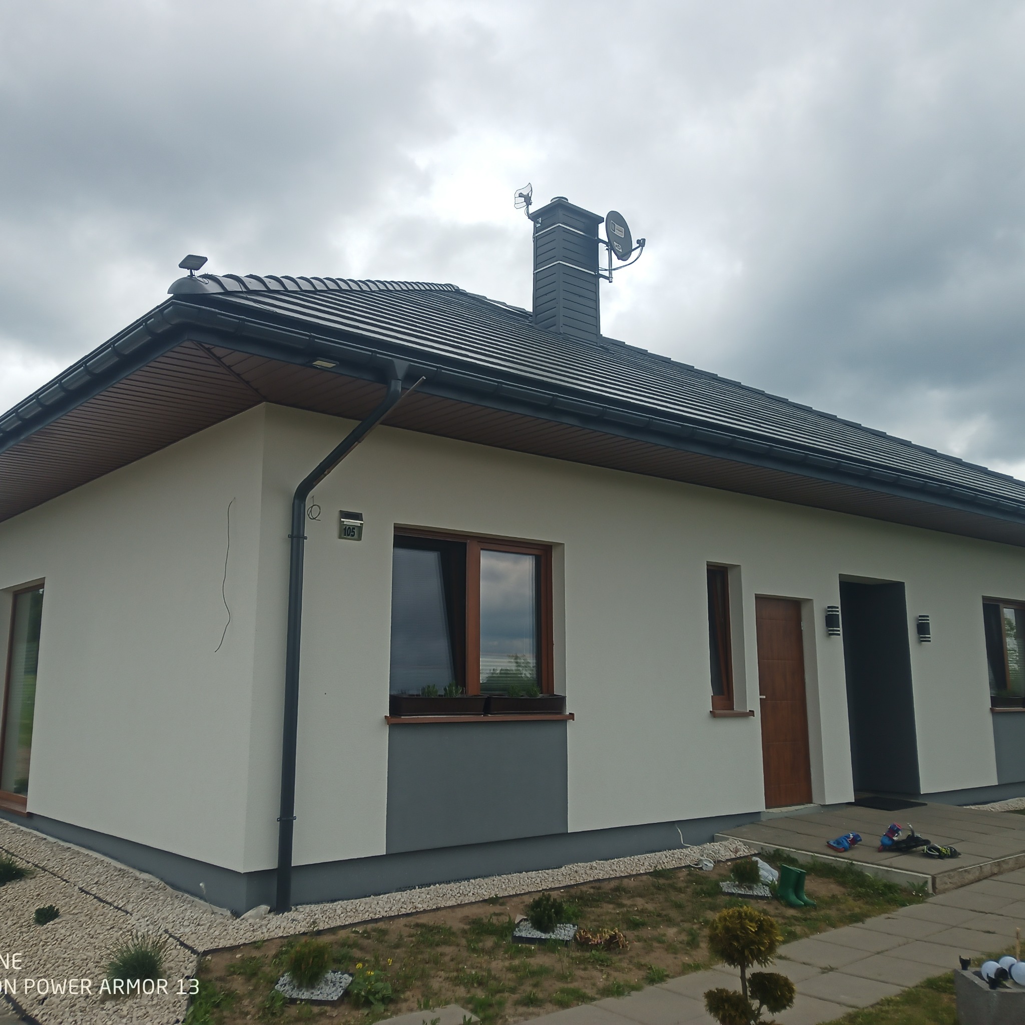 Jednopiętrowy dom z jasną elewacją, ciemną dachówką i szarymi akcentami, widoczny komin z antenami satelitarnymi, otoczony kamieniami i zielenią, z butami i zabawkami przed wejściem.