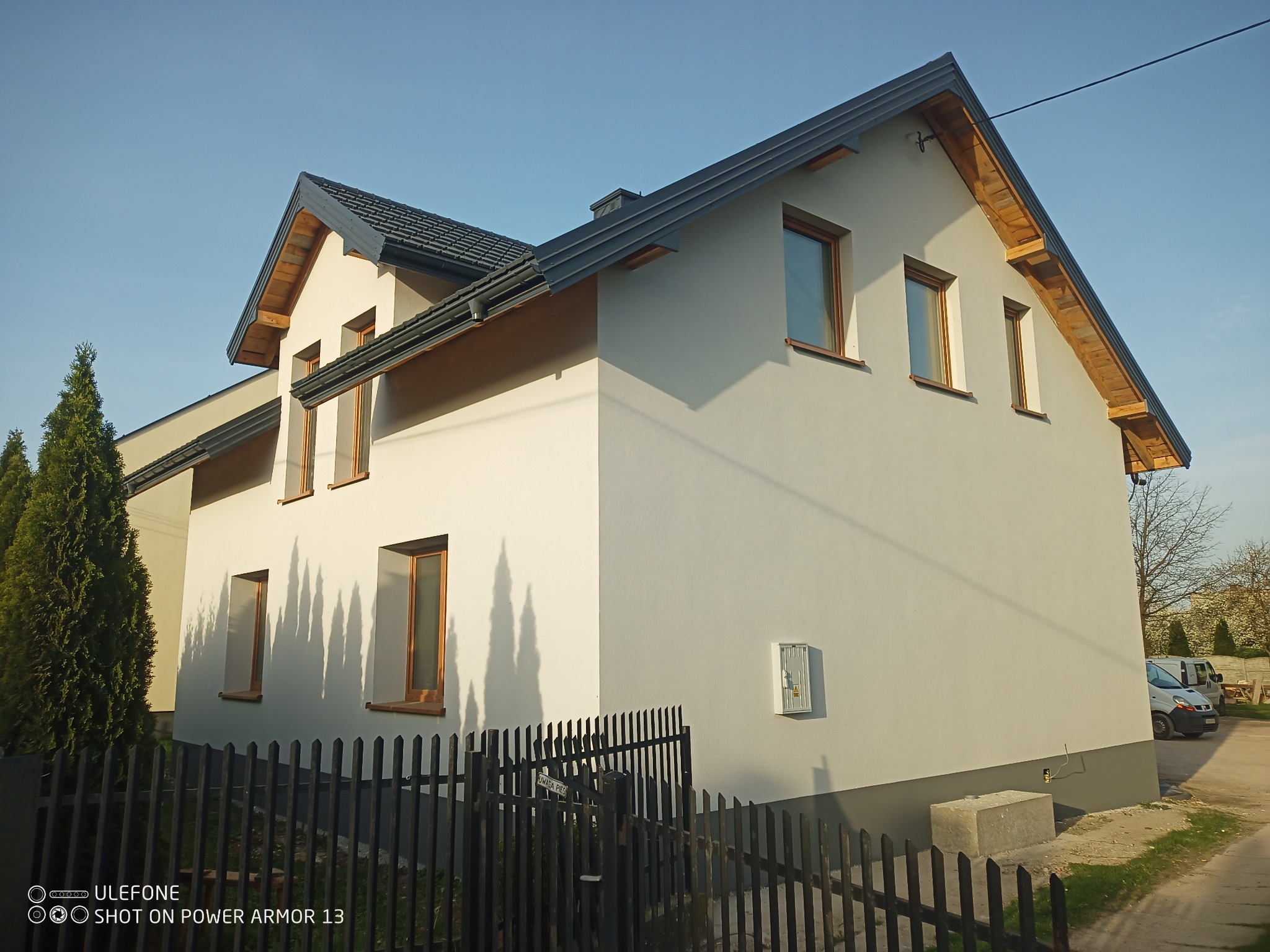 Nowoczesny, dwukondygnacyjny dom jednorodzinny z ciemną dachówką i jasnoszarą elewacją, widziany zza czarnego, metalowego ogrodzenia w słoneczny dzień.