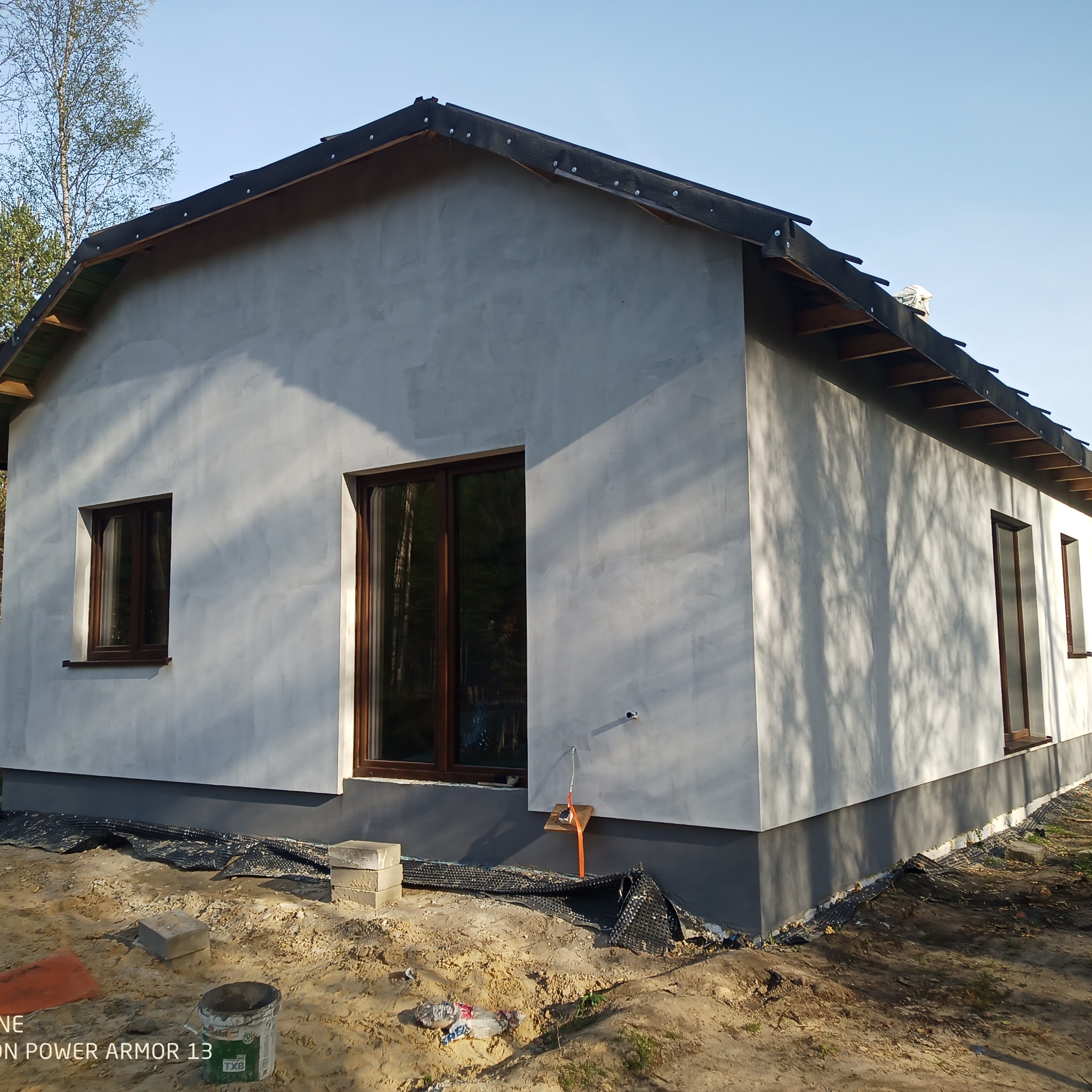 Dom jednorodzinny po nałożeniu szarego tynku elewacyjnego, widoczne brązowe ramy okienne, przygotowany teren wokół budynku z materiałami budowlanymi i pustakami.