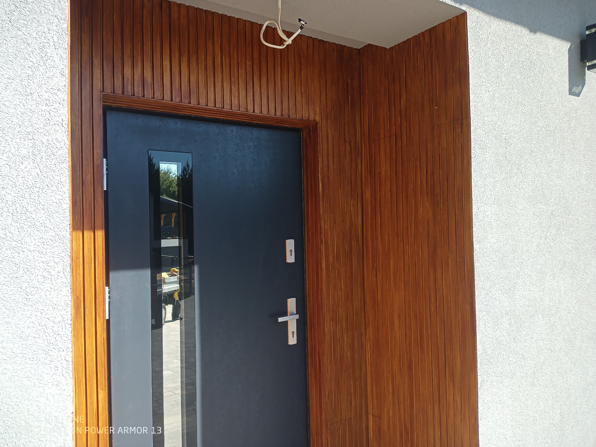 Wejście do domu z ciemnymi drzwiami i drewnianą boazerią, widoczne nierówności faktury tynku na ścianie.