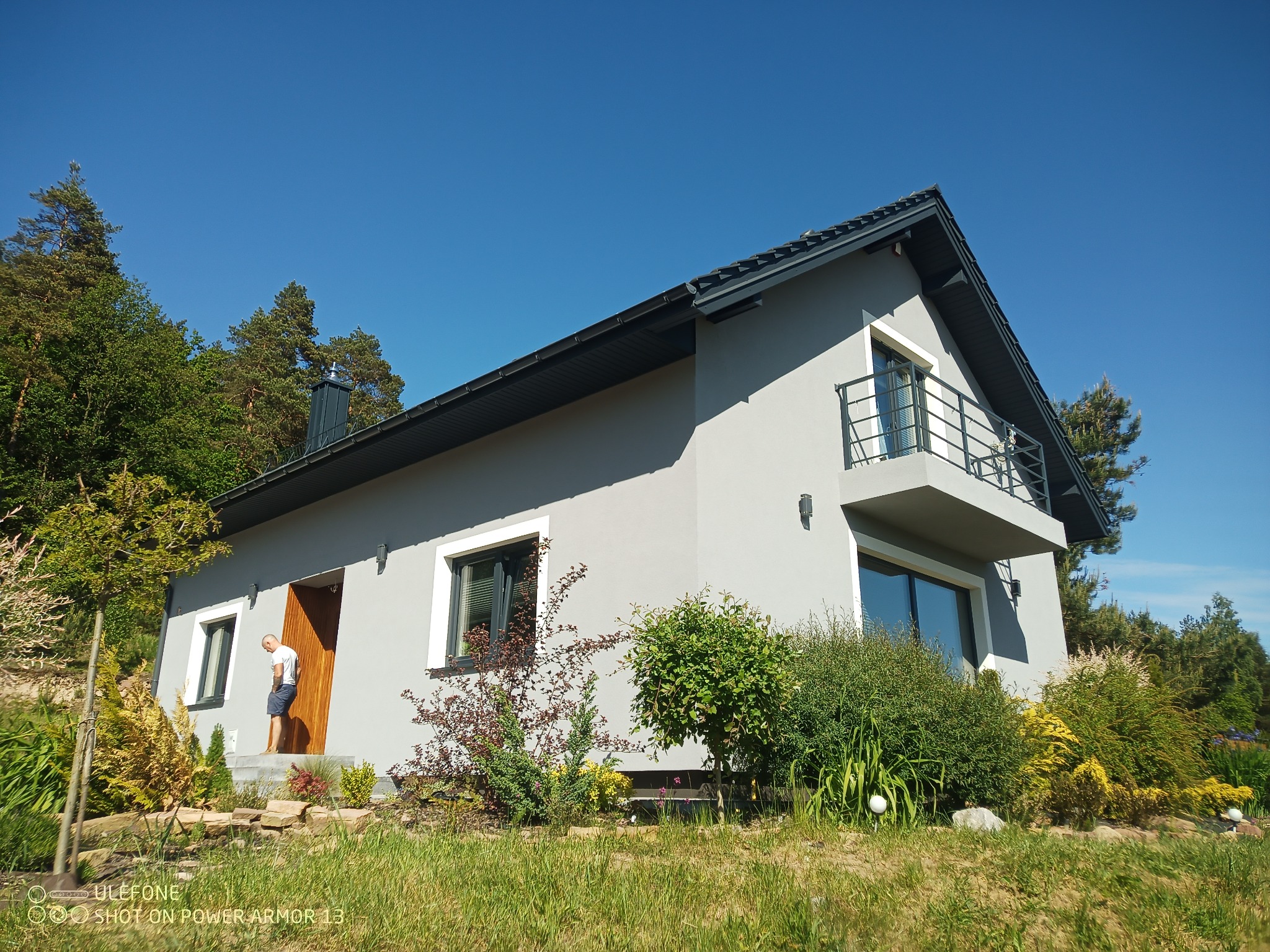 Nowoczesny dom jednorodzinny z szarą elewacją, balkonem z metalową balustradą i drewnianymi drzwiami wejściowymi, otoczony zielenią i drzewami w słoneczny dzień.