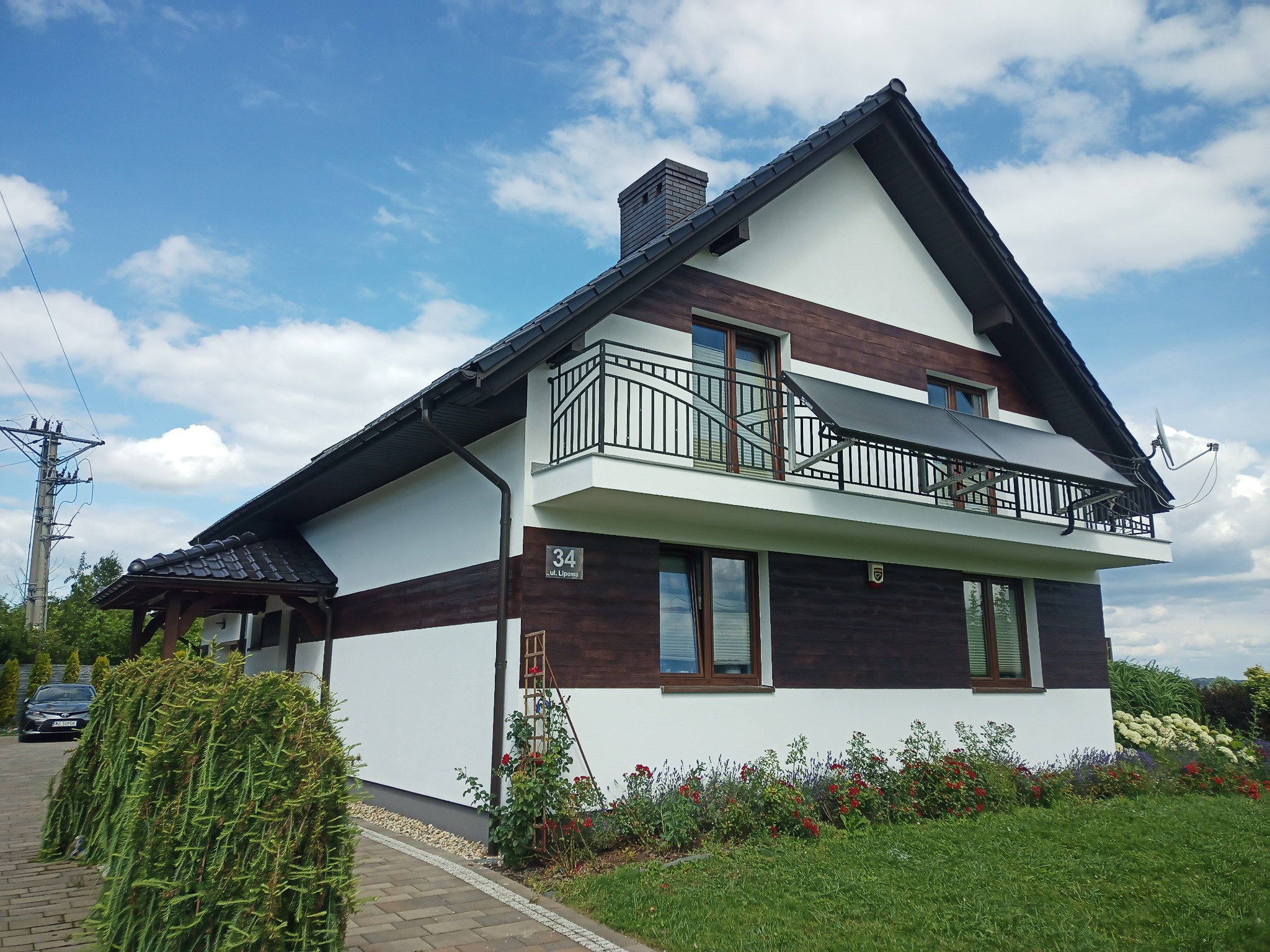 Dwukondygnacyjny dom z balkonem, elewacją w kolorze białym i brązowym, czarnym dachem i panelami słonecznymi na balkonie, otoczony zielenią i kwiatami w słoneczny dzień.