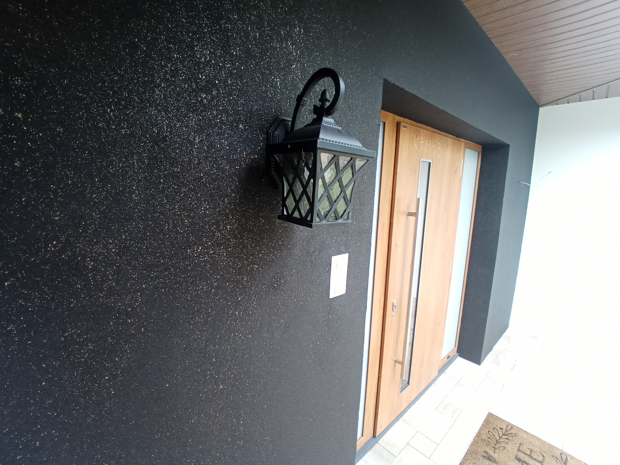 Elewacja budynku w kolorze ciemnego grafitu ze złotymi drobinkami, oświetlona ozdobnym, czarnym kinkietem w stylu retro, obok drewniane drzwi wejściowe z przeszkleniem.