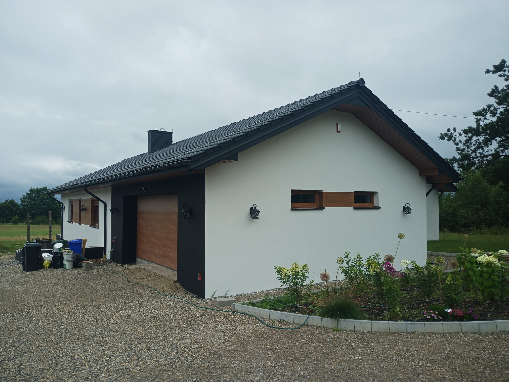 Nowoczesny dom jednorodzinny z białą elewacją, czarnymi elementami wykończeniowymi i drewnianą bramą garażową, otoczony żwirowym podjazdem i rabatą kwiatową. Widoczne lampy zewnętrzne w stylu retro...