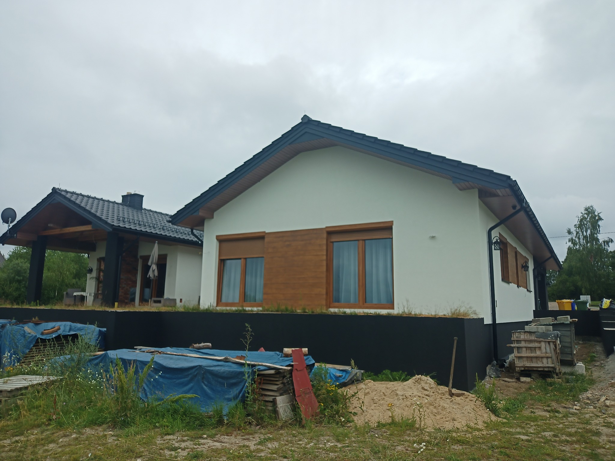 Nowoczesny dom jednorodzinny z białymi ścianami i drewnianymi akcentami okiennymi, widoczny z poziomu gruntu, z elementami placu budowy w otoczeniu.