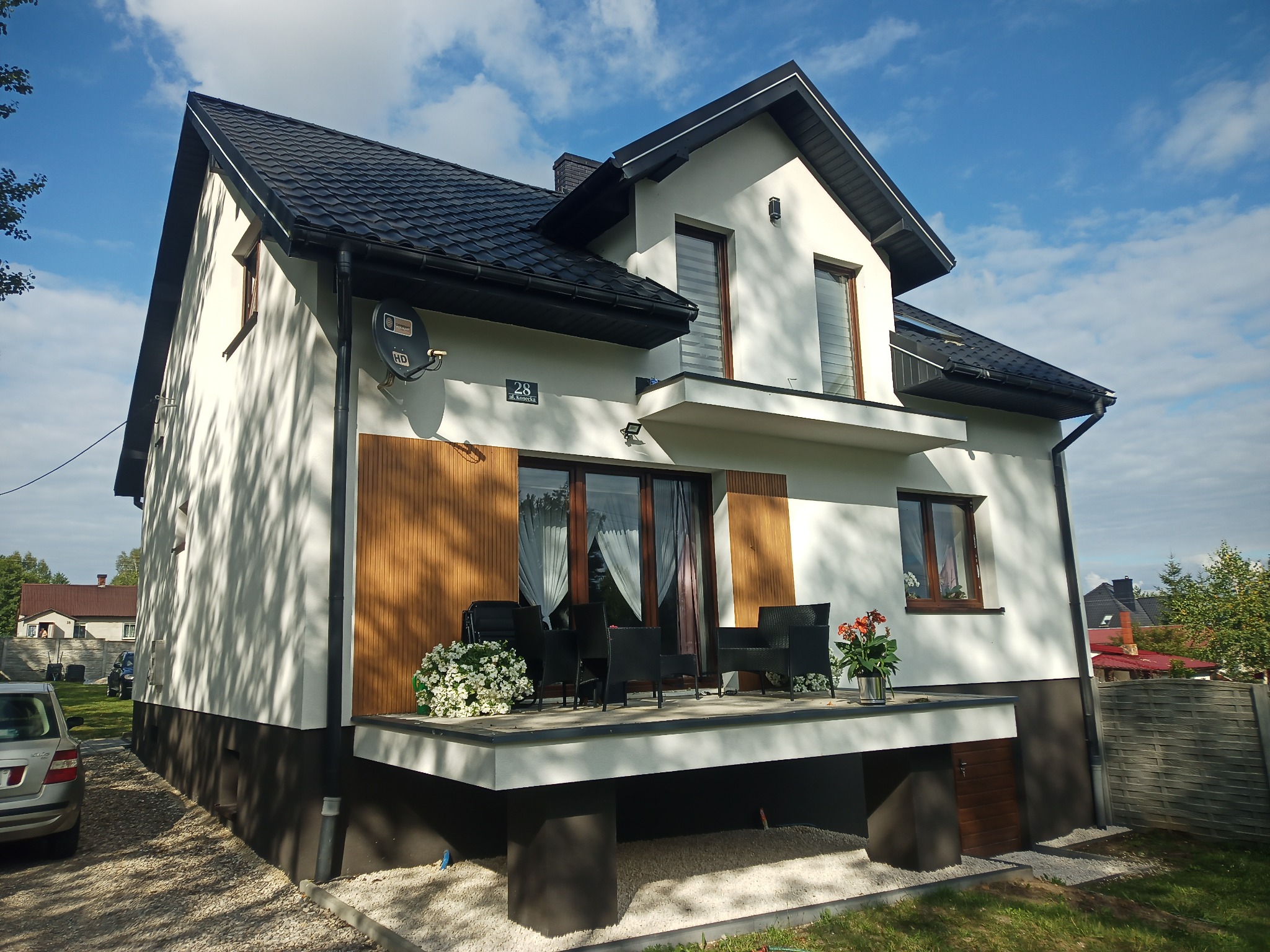 Nowoczesny, dwukondygnacyjny dom z białymi ścianami, ciemnym dachem i drewnianymi akcentami, z tarasem wyposażonym w czarne meble ogrodowe i kwiaty, widok z boku na tle błękitnego nieba z chmurami.