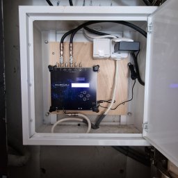 Digi-Media Paweł Kopczyński - Wnętrze skrzynki z zamontowanym wzmacniaczem sygnału antenowego Telmor Alpha+ z podłączonymi kablami koncentrycznymi i zasilaniem, widoczne złącza antenowe i gniazdo USB.