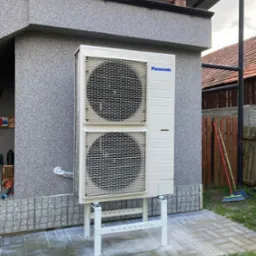 Biała, dwusekcyjna pompa ciepła marki Panasonic zamontowana na metalowym stelażu przy ścianie budynku z szarą elewacją i drewnianą konstrukcją dachu.