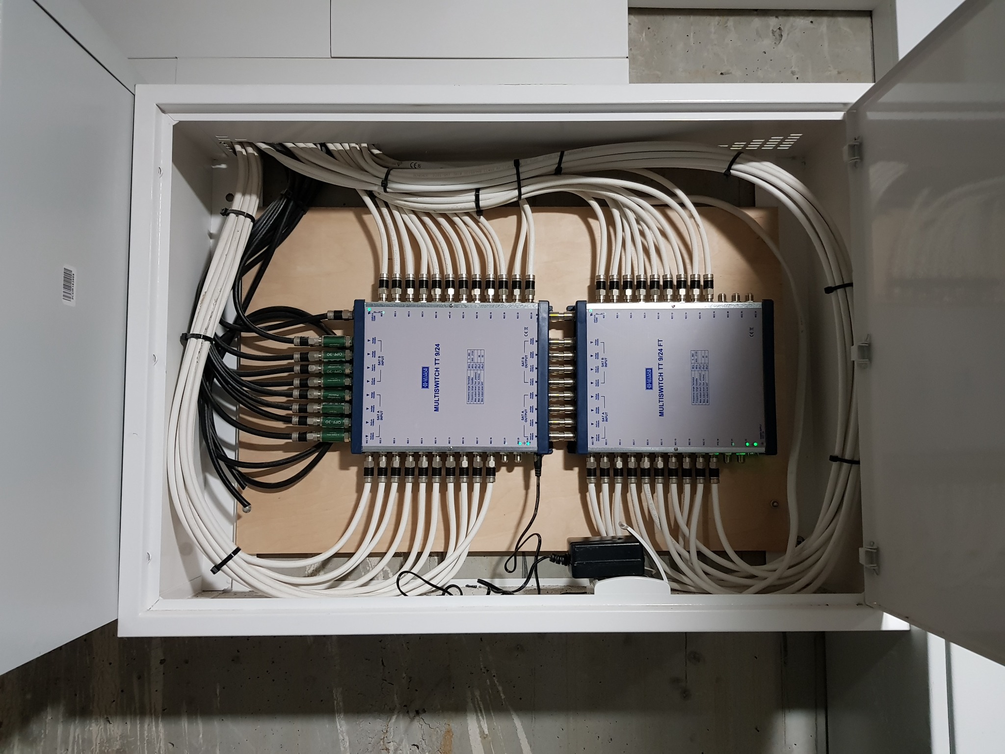 Rozdzielnica z dwoma multiswitchami TT 9/24, liczne kable koncentryczne uporządkowane opaskami zaciskowymi, zamontowane w białej szafce na drewnianej płycie, widoczne zielone diody LED.
